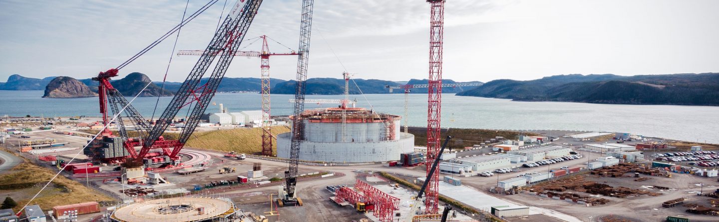 Large construction site near a body of water with several cranes, a cylindrical industrial structure, scattered building materials, and distant hills under a partly cloudy sky.