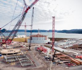 Large construction site near a body of water with several cranes, a cylindrical industrial structure, scattered building materials, and distant hills under a partly cloudy sky.