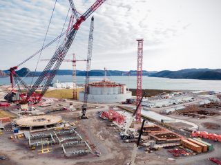 Large construction site near a body of water with several cranes, a cylindrical industrial structure, scattered building materials, and distant hills under a partly cloudy sky.