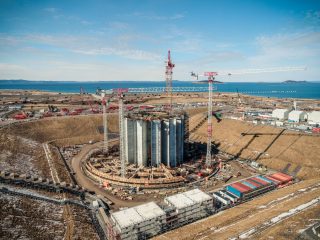 A construction site with several tall cylindrical structures surrounded by cranes and scaffolding, set in a large open landscape near water under a blue sky. Temporary buildings and shipping containers are nearby.