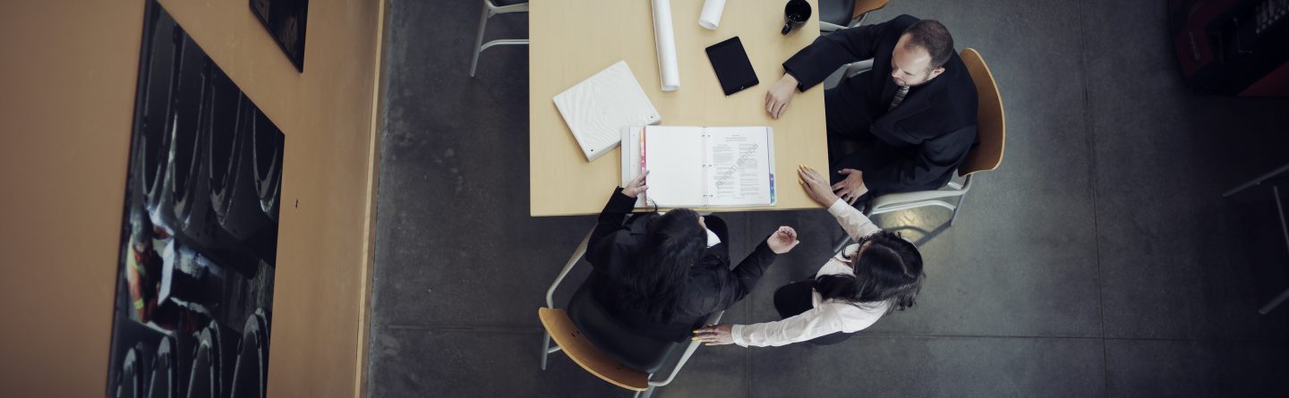 Aerial view of three people in business attire sitting around a rectangular table with documents, a tablet, coffee mug, and rolled-up papers in a modern office setting.