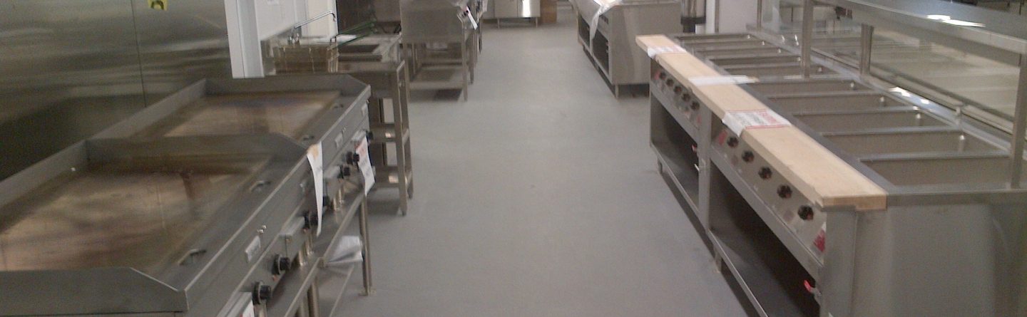A commercial kitchen with stainless steel appliances, including griddles, ovens, and food preparation counters. The space is clean and well-organized, with equipment covered or labeled, and bright fluorescent lighting overhead.