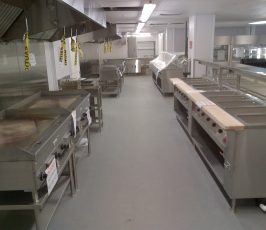 A commercial kitchen with stainless steel appliances, including griddles, ovens, and food preparation counters. The space is clean and well-organized, with equipment covered or labeled, and bright fluorescent lighting overhead.