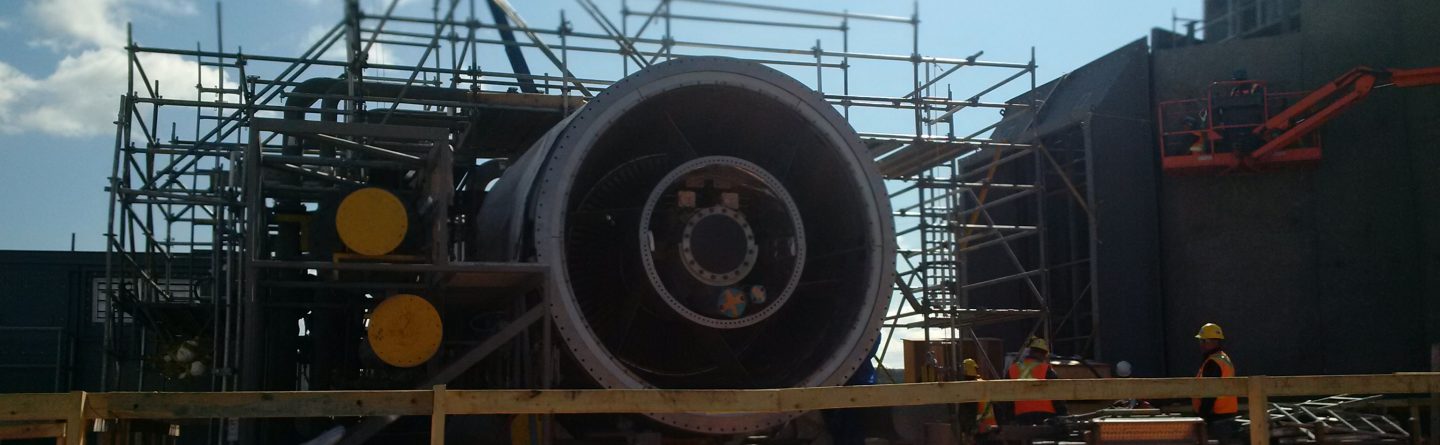 A large jet engine is surrounded by scaffolding at an industrial construction site, with workers in safety gear nearby and bright sunlight casting shadows.