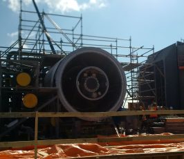 A large jet engine is surrounded by scaffolding at an industrial construction site, with workers in safety gear nearby and bright sunlight casting shadows.