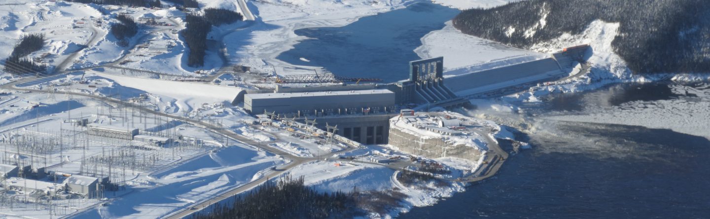 Aerial view of a large hydroelectric dam surrounded by snow and ice, with a river flowing through a wintry landscape and mountains in the background.