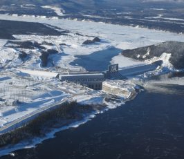 Aerial view of a large hydroelectric dam surrounded by snow and ice, with a river flowing through a wintry landscape and mountains in the background.