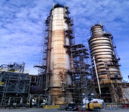 Two tall industrial towers surrounded by scaffolding and machinery at a refinery or chemical plant, under a blue sky with scattered clouds.