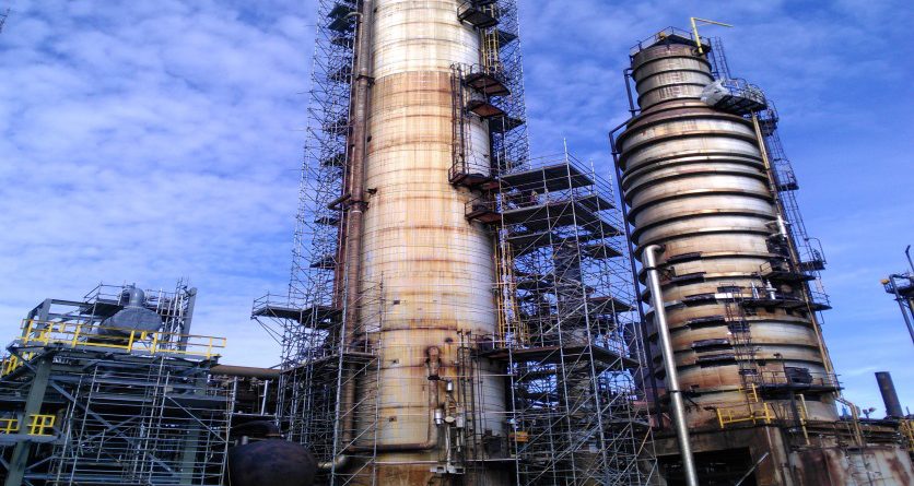 Two tall industrial towers surrounded by scaffolding and machinery at a refinery or chemical plant, under a blue sky with scattered clouds.
