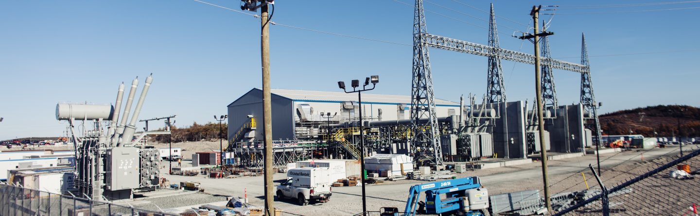 An electrical substation with metal towers, transformers, utility poles, and construction equipment. Several vehicles and electrical components are inside a fenced area under a clear blue sky.