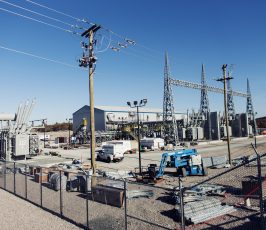 An electrical substation with metal towers, transformers, utility poles, and construction equipment. Several vehicles and electrical components are inside a fenced area under a clear blue sky.
