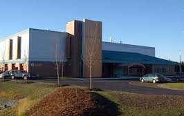A modern, single-story building with a flat roof and large windows, surrounded by a few parked cars, small leafless trees, and a paved driveway under a clear blue sky.