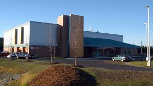 A modern, single-story building with a flat roof and large windows, surrounded by a few parked cars, small leafless trees, and a paved driveway under a clear blue sky.