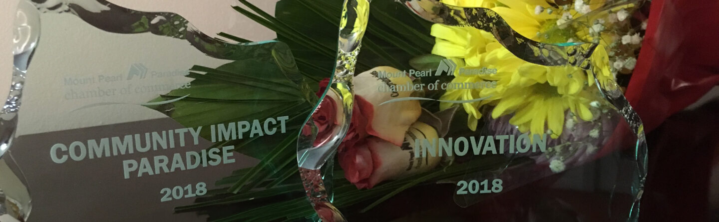 Two glass awards labeled "COMMUNITY IMPACT PARADISE 2018" and "INNOVATION 2018" sit side by side in front of a bouquet of yellow, red, and white flowers.