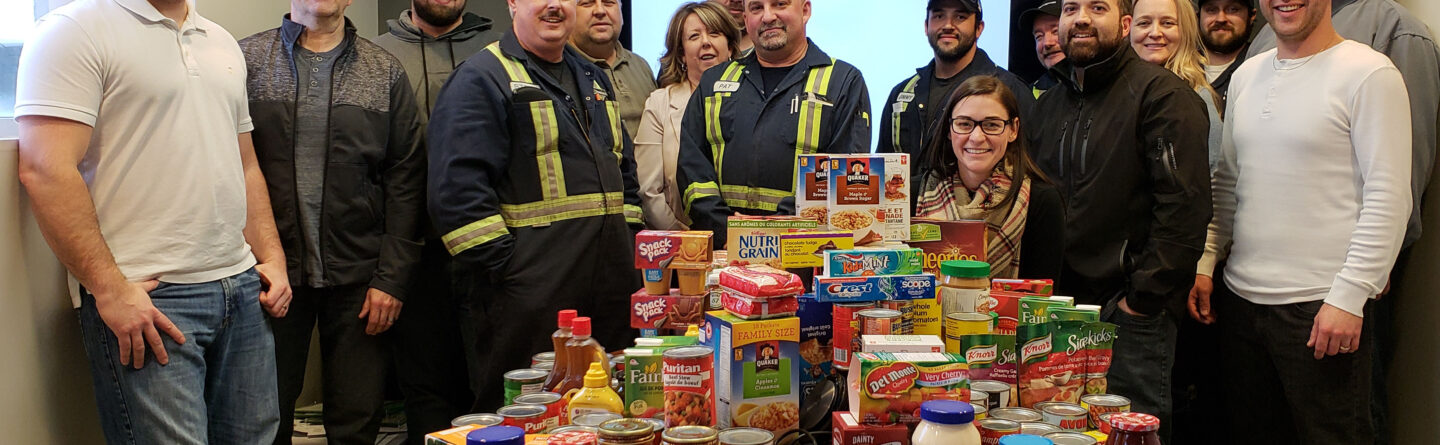 A group of people stand smiling around a table filled with food donations, including canned goods, pasta, and cereal boxes, in an office setting. A company logo is displayed on a screen in the background.