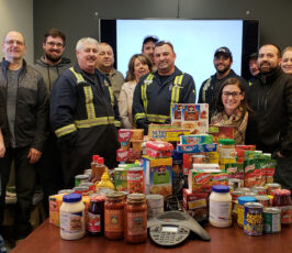 A group of people stand smiling around a table filled with food donations, including canned goods, pasta, and cereal boxes, in an office setting. A company logo is displayed on a screen in the background.