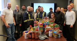 A group of people stand smiling around a table filled with food donations, including canned goods, pasta, and cereal boxes, in an office setting. A company logo is displayed on a screen in the background.