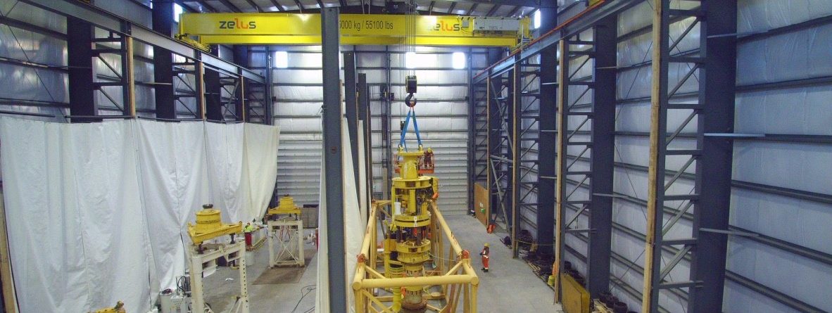 A large industrial warehouse with yellow overhead cranes, workers in safety gear, and heavy machinery in the center. The cranes are labeled "zelus" and "25000 kg / 55,000 lbs." The space is well-lit and organized.