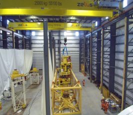 A large industrial warehouse with yellow overhead cranes, workers in safety gear, and heavy machinery in the center. The cranes are labeled "zelus" and "25000 kg / 55,000 lbs." The space is well-lit and organized.