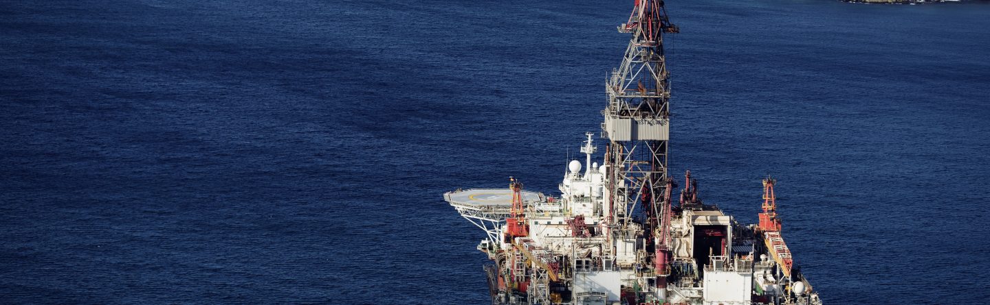 An offshore oil rig sits in deep blue water near a coastline with rocky terrain and scattered houses under a partly cloudy sky.