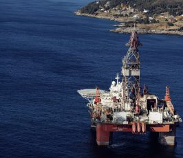 An offshore oil rig sits in deep blue water near a coastline with rocky terrain and scattered houses under a partly cloudy sky.