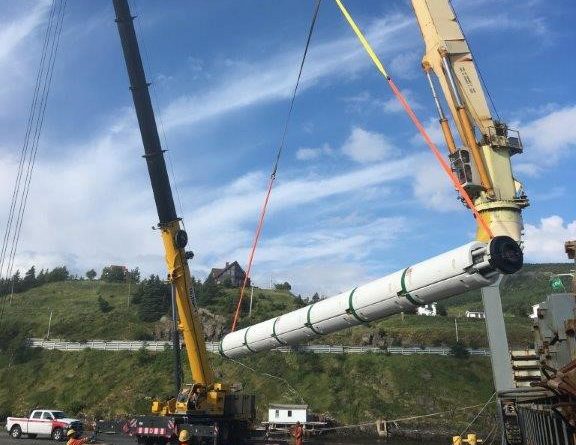 Two large cranes lift a long cylindrical object using straps at a construction site. Workers in safety gear stand nearby, and a hillside with buildings and a white truck are visible in the background.