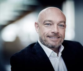 A middle-aged man with a shaved head and trimmed beard smiles while wearing a dark suit jacket and white shirt, seated in a modern, blurred office setting.