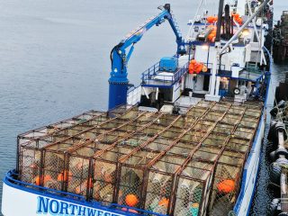 A large fishing boat named NORTHWESTERN, loaded with crab pots and orange buoys, is docked at a pier in a cold, coastal area with snow-covered hills in the background.