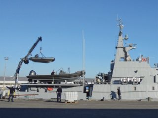 A naval ship docked at a port with crew members nearby. A crane on the ship is lifting a small boat onto the deck under a clear blue sky.