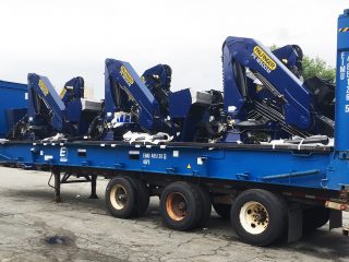 A flatbed trailer loaded with several large blue Palfinger cranes, secured and ready for transport, is parked on a paved lot. The cranes are folded and have visible yellow branding.