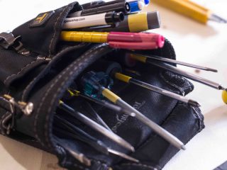 A close-up of a black tool pouch filled with various pens, screwdrivers, and a utility knife, resting on a white surface.