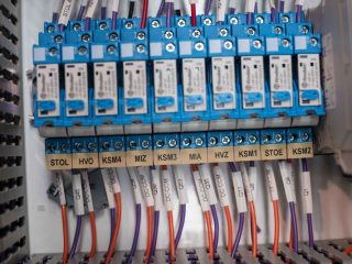 A close-up view of a control panel showing blue electrical relays with labeled wires connected below, each marked with codes such as STOL, HVO, KSM4, MIZ, KSM3, MIA, HVI, STOE, and KSM2.