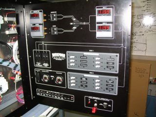 Black control panel with digital displays showing 11.99 volts, various switches, dials, buttons, and labeled sections for BUS 1 and BUS 2, with wiring diagrams and labels. Whiteboard and cardboard box in background.
