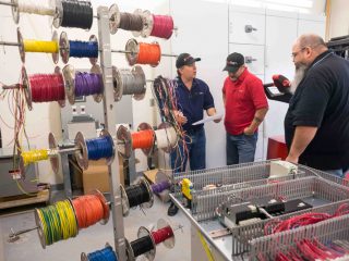 Three men stand in a workshop discussing wiring. Multiple spools of colored wire are mounted on a rack, and an open control panel with electronics and wires is visible in the foreground.