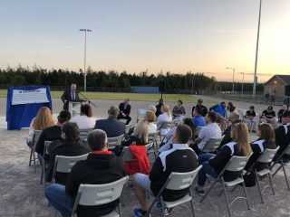 A group of people sit on folding chairs outdoors at sunset, listening to a man speaking at a podium. A blue-covered sign labeled 