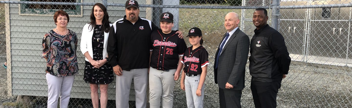 Seven people, including two boys in baseball uniforms and five adults in business or casual attire, stand together on a baseball field near a chain-link fence and a small building, smiling at the camera.