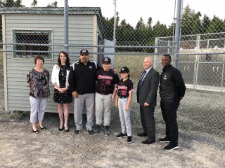 Seven people, including two boys in baseball uniforms and five adults in business or casual attire, stand together on a baseball field near a chain-link fence and a small building, smiling at the camera.