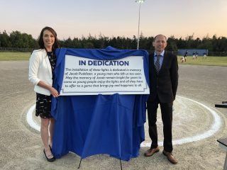 Two people stand on a baseball field holding a sign that dedicates the new field lights to the memory of Jacob Dubéister, with a youth baseball game and trees visible in the background at sunset.