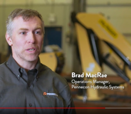 A man in a grey Pennecon shirt is speaking in an industrial setting with machinery in the background. On-screen text reads: "Brad MacRae, Operations Manager, Pennecon Hydraulic Systems.