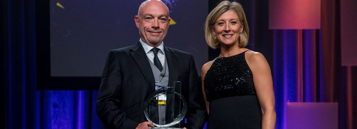 A man in a suit holding an award stands beside a woman in a black dress. Behind them is a screen displaying the name "Larry Puddister" in bold letters, with stage lighting and geometric decor in the background.