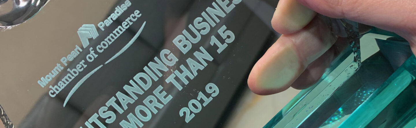 A hand holds a glass award with text reading "Outstanding Business More Than 15, 2019, Pennco Tech" from the chamber of commerce. The award has a blue base and jagged edges, with an office background visible.