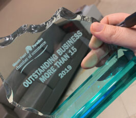 A hand holds a glass award with text reading "Outstanding Business More Than 15, 2019, Pennco Tech" from the chamber of commerce. The award has a blue base and jagged edges, with an office background visible.
