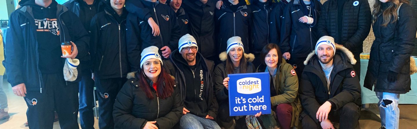 A group of people dressed warmly in winter coats and hats pose indoors in front of blue and yellow decorations. One person holds a sign reading "Coldest Night - it