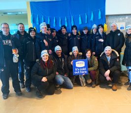 A group of people dressed warmly in winter coats and hats pose indoors in front of blue and yellow decorations. One person holds a sign reading "Coldest Night - it
