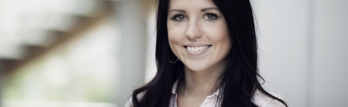 A woman with long dark hair, wearing a light-colored button-up shirt, smiles at the camera in a softly lit indoor setting with a blurred background.