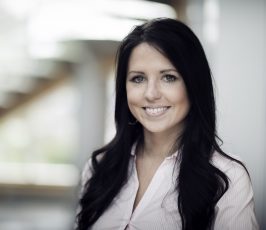 A woman with long dark hair, wearing a light-colored button-up shirt, smiles at the camera in a softly lit indoor setting with a blurred background.