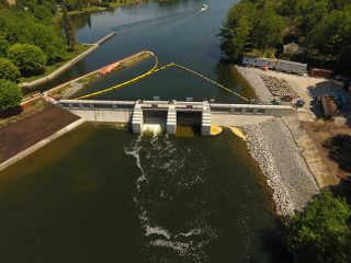 Aerial view of a dam with three gates releasing water into a river. Surrounding the dam are trees, a boat on the water, a construction area, and a shoreline with rocks and grass.