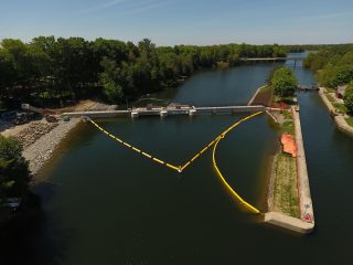 Aerial view of a river with a dam and yellow floating barriers forming a V shape in the water, surrounded by trees and construction equipment on the shore.
