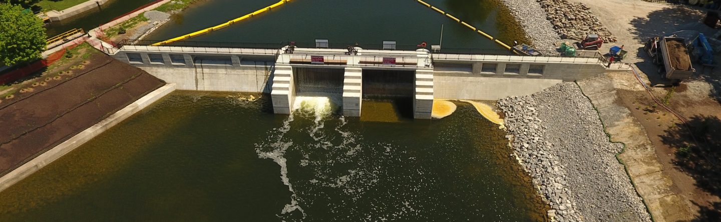 Aerial view of a small dam and spillway on a river, with water flowing through gates, surrounded by trees, construction equipment, and a boat visible in the background on the water.