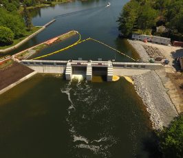Aerial view of a small dam and spillway on a river, with water flowing through gates, surrounded by trees, construction equipment, and a boat visible in the background on the water.